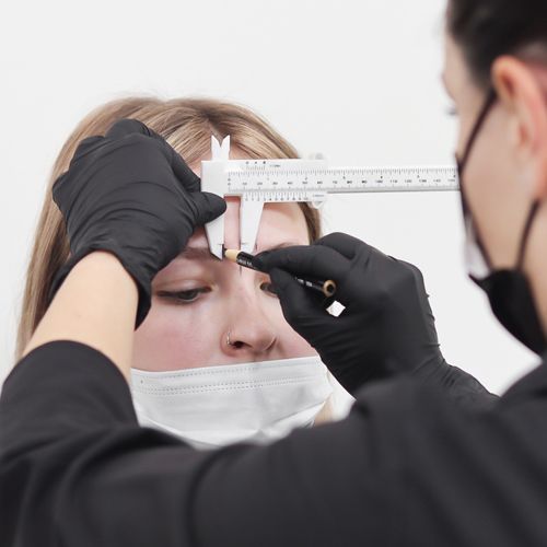 Esthetician using measuring client's eyebrows while using black pencil to mark brow shape.