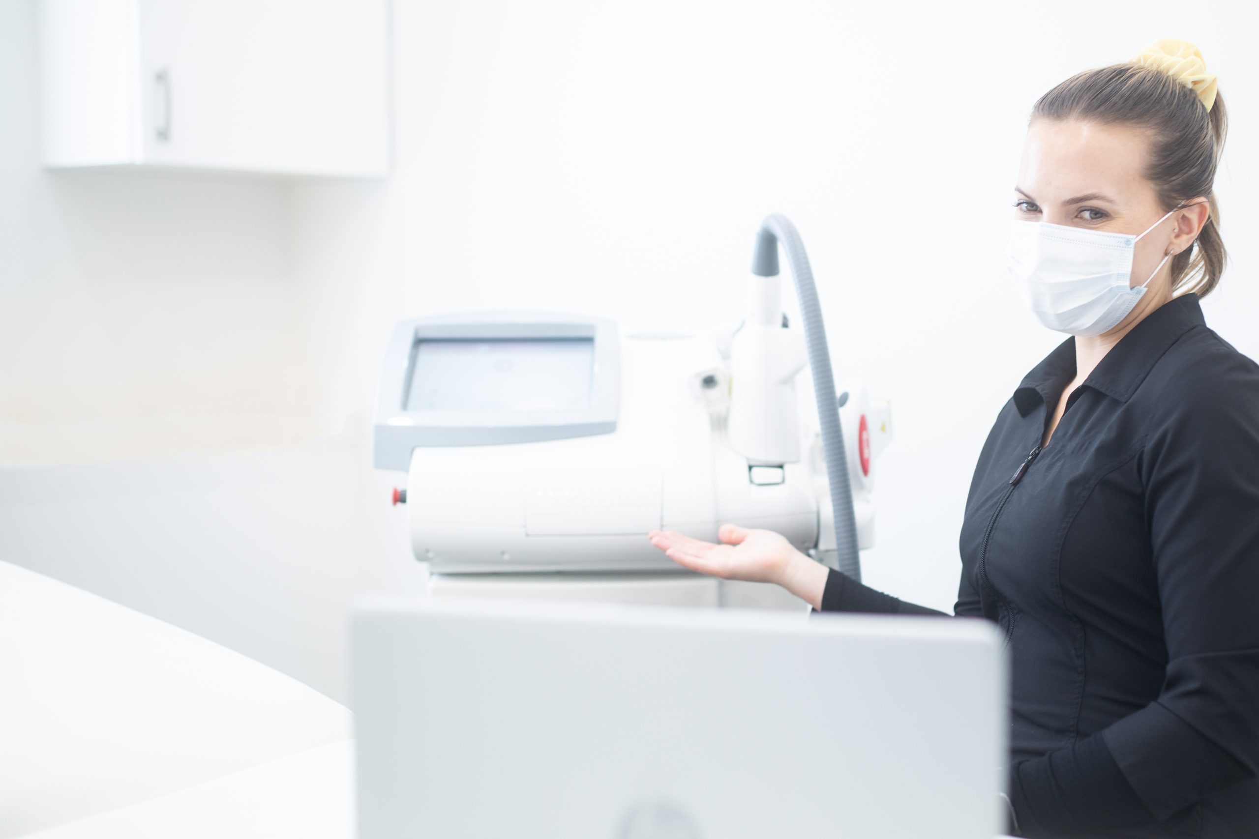 Female esthetician wearing black professional smock and white face mask using hand to gesture towards laser hair removal machine.