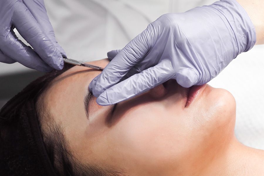 gloved hands holding dermaplaning scalpel against woman's forehead, removing vellus hairs