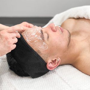 Male client with eyes closed and black headband laying face up with exfoliating skin care product on face.
