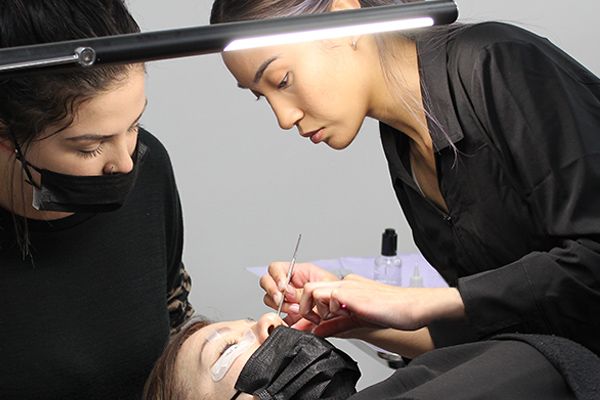 Female lash extension educator in black smoke applying lash extension to client's eyes with tweezers. Client is laying on their back with eyes closed. Student dressed in black attire and black face mask watches educator's demonstration.