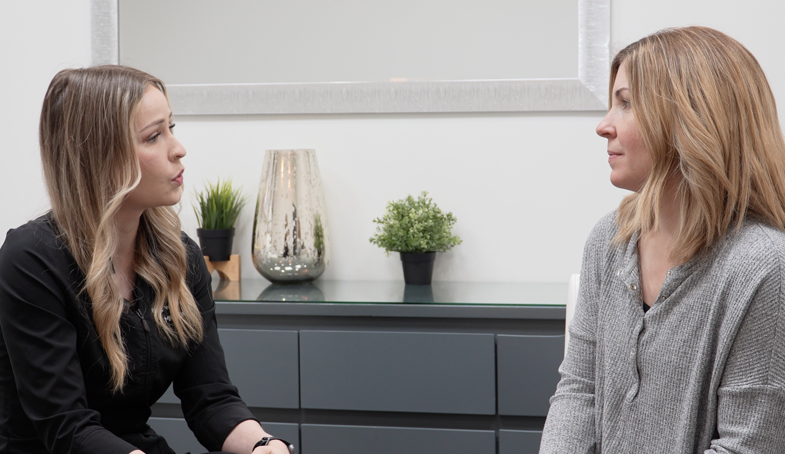 "esthetician in a black smock talking to a client in a grey sweater"
